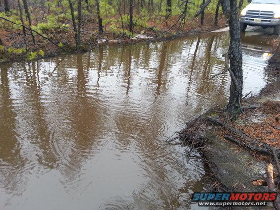 White SUV driving through a muddy water crossing in a wooded area