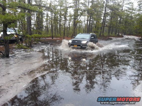 Dark Toyota 4Runner splashing through a muddy puddle on a dirt road, another white SUV visible behind it