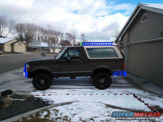 Dark gray Ford Bronco with a tan top, parked in a driveway with snow on the ground. It has blue aftermarket bumpers and a roof rack
