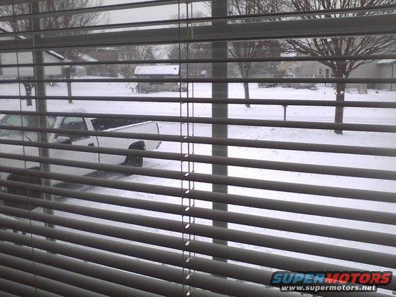 White pickup truck partially obscured by blinds, parked on a snow-covered street