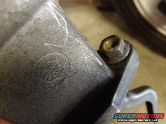 Close-up of a metal automotive part showing a bolt and nut, with some scratch marks and a circular embossed symbol