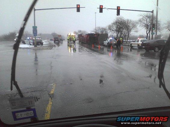Accident scene with police cars, overturned truck, and emergency personnel on a wet road