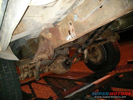 Underside of a heavily rusted vehicle frame with drivetrain components and a tire visible