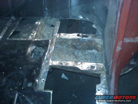 Red vehicle interior floor pan with rust and cut-out sections, showing metal framing