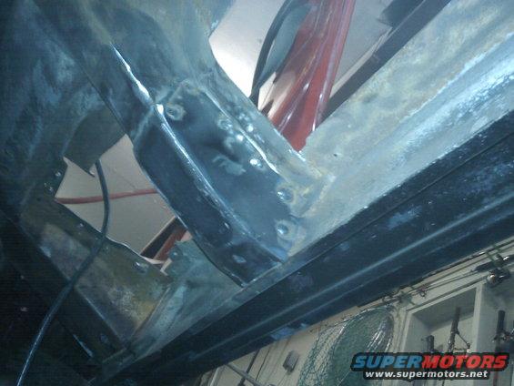 Underside of a vehicle frame with visible welds and rust. Red paneling is visible in the background