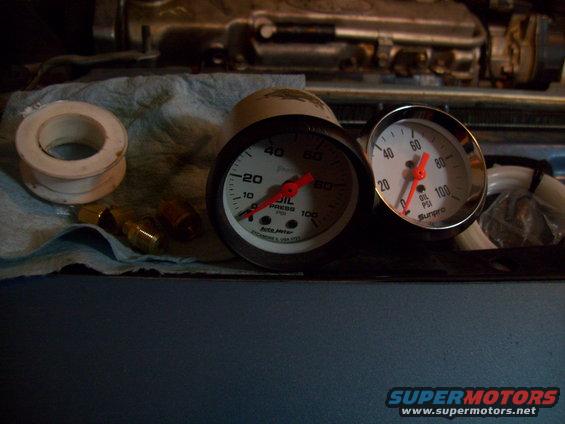 Two oil pressure gauges and brass fittings on a blue surface, with engine parts in the background