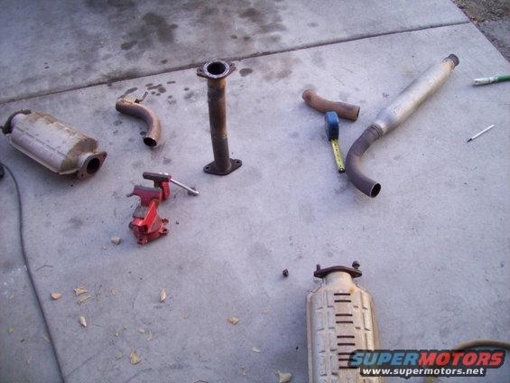 Exhaust pipes, catalytic converter, and muffler laid out on concrete with a vise and tape measure