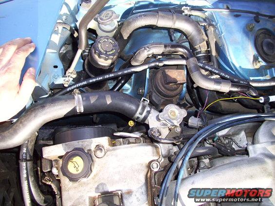 Blue car engine bay with hoses, fluid reservoirs, and engine components visible. A hand is in the upper left