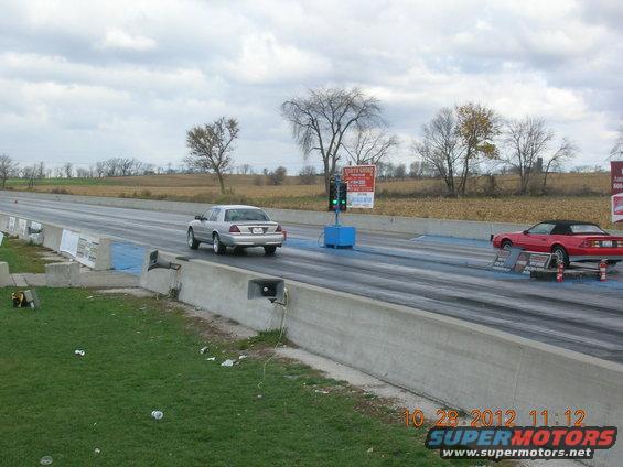 Silver sedan and red convertible on drag strip, trees and fields in background