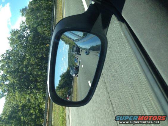 View in side mirror shows cars on a highway with trees and blue sky