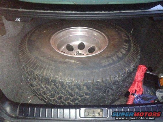 Spare tire with aluminum rim in a car trunk, next to a red emergency kit