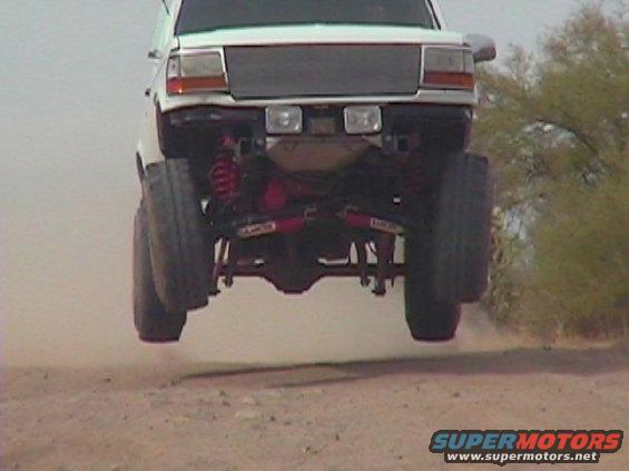 White pickup truck mid-air jump, showing lifted suspension and large tires. The grille and headlights are visible