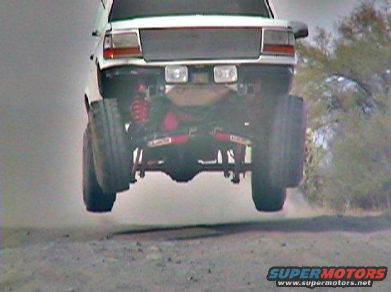 White SUV with lifted suspension airborne, rear view. Red shocks visible