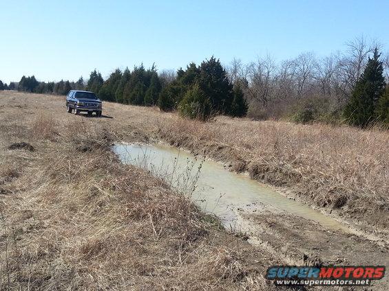 Blue Ford Bronco driving on a dirt path next to a muddy ditch