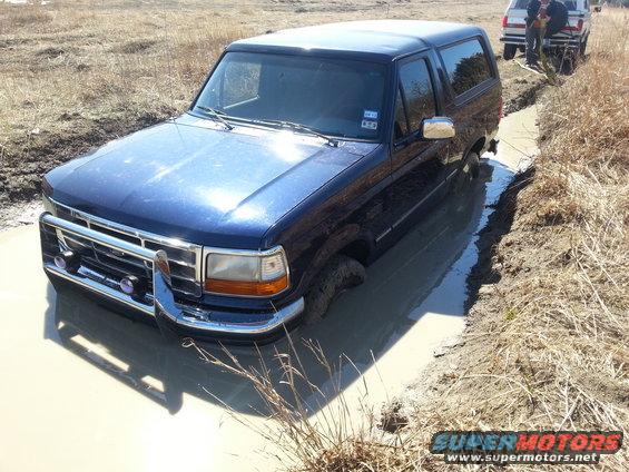 Blue SUV stuck in a muddy ditch, another vehicle and people visible in background