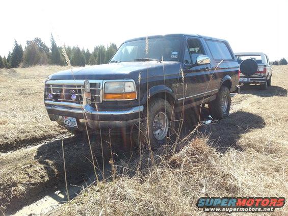 Dark blue Ford Bronco stuck in mud, front quarter view. Another white truck is behind it