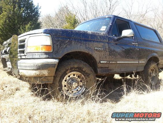 Muddy blue Ford Bronco parked in dry grass. Bronco badge visible on fender