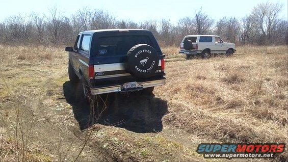 Dark blue Ford Bronco on a dirt track, white Ford Bronco in grassy field behind it