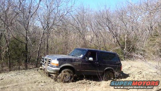 Dark blue Ford Bronco stuck in mud, front grille visible, trees in background