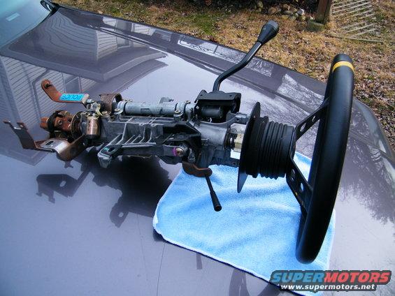 Steering column assembly with shifter lever and aftermarket steering wheel, resting on a blue cloth on a car hood. A blue 'POLICE' tag is visible