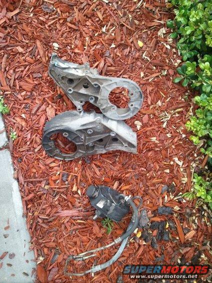 Two metal brackets and a black power steering pump with hoses lie on red mulch