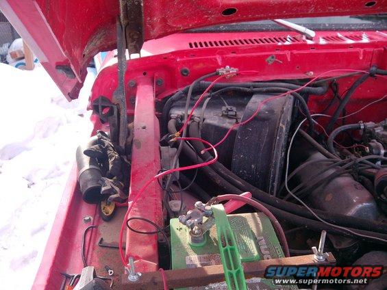 Red engine bay with visible battery, wiring, and engine components. A pair of gloves rests near a black hose