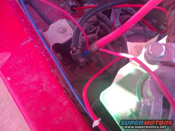 close-up of red vehicle's battery terminals and wiring, with some text visible on a piece of paper