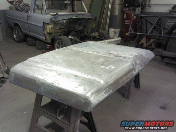 Bare metal truck hood on a stand in a workshop, with a disassembled truck in the background