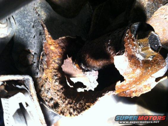 Close-up of heavily rusted metal, likely a vehicle frame, with a large hole showing severe corrosion. A bolt is visible