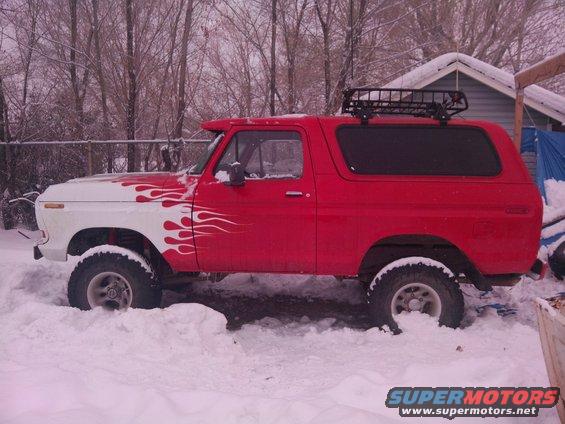 Red and white Ford Bronco with a roof rack, partially covered in snow, seen from the side