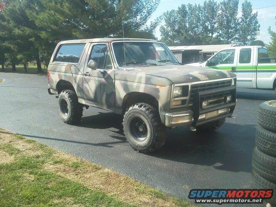 Camouflaged Ford Bronco parked on asphalt, with a white pickup truck in the background
