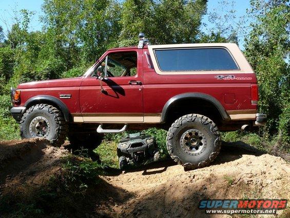 Red Ford Bronco with tan top and off-road tires on a dirt trail, with an ATV underneath