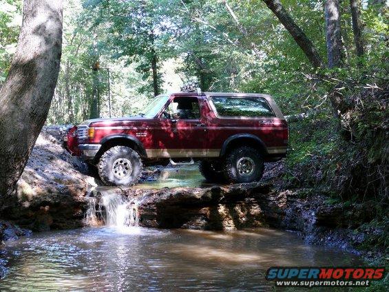 Red Ford Bronco with off-road tires driving through a stream in a wooded area