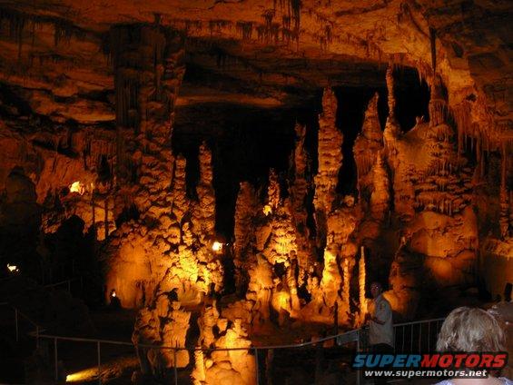 Cave interior with illuminated stalactites and stalagmites, a railing, and two people visible