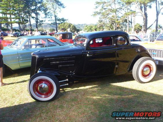 Black 1934 Ford hot rod coupe with red interior and wide whitewall tires. Another car is parked to its left