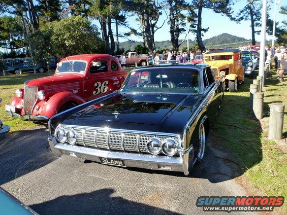 Front view of a black 1963 Lincoln Continental with a custom grille and license plate that says SLAMM. Red car with number 36 is to the left