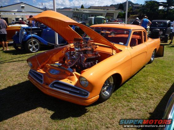 Orange custom hot rod with hood open showing V8 engine. Another blue hot rod is visible behind it