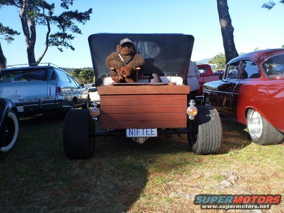 Rear view of a custom hot rod with a wooden bed, large rear tires, and a teddy bear wearing a hat and sunglasses in the driver's seat. License plate reads 'NIFTEE'. A red classic car is parked to the right