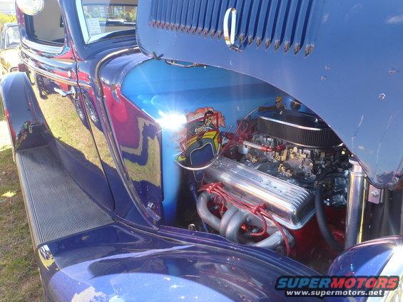 Blue hot rod with custom painted mural on inner fender and chrome V8 engine. Headers and air intake visible