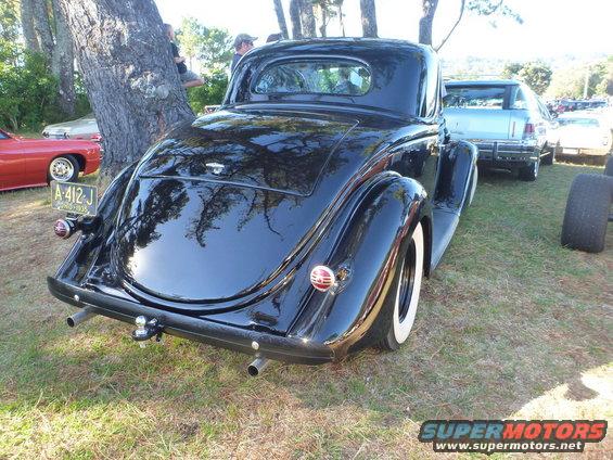 Black coupe with whitewall tires and a trailer hitch, parked on grass. Other cars visible in background