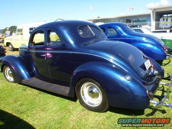 Blue hot rod coupe parked on grass, another blue car behind it. Yellow trailer and building in background