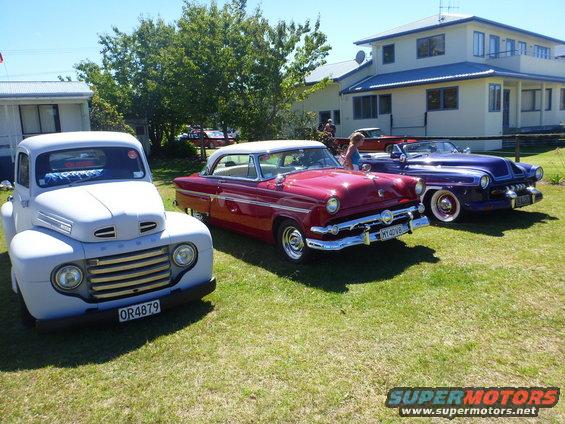 Three classic cars parked on grass: a light-colored truck, a red coupe, and a purple convertible