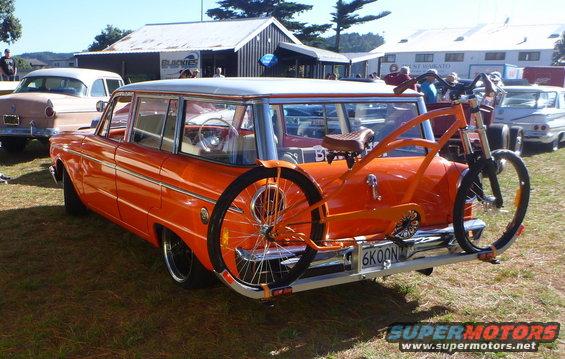 Orange 1959 Ford Country Sedan station wagon with a custom bicycle mounted on the rear rack. License plate reads 6KOON7