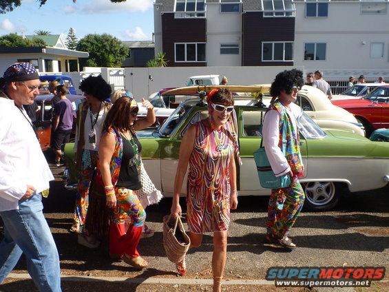 Green and white 1958 car with surfboard on roof, people in retro outfits walking by