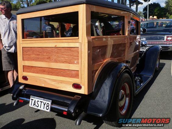 Rear view of a custom woodie wagon with a 'TASTY8' license plate. Another car is visible to the right
