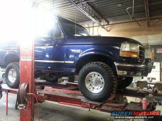 Blue Ford Bronco on a car lift in a garage. The Bronco has a chrome bumper and large tires