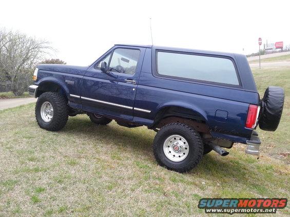 Blue Ford Bronco parked on a grassy hill, showing the side and rear with a spare tire mounted on the back