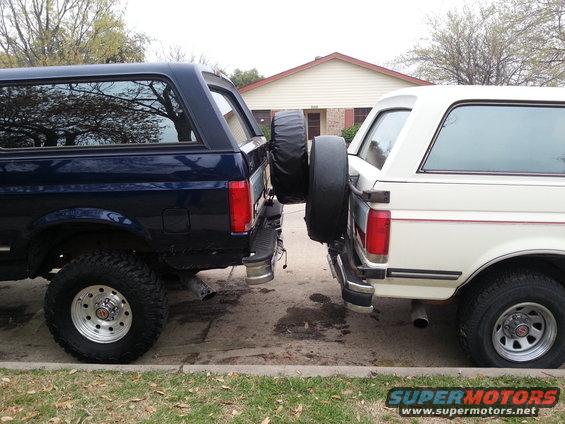 Two Ford Bronco SUVs parked bumper to bumper, one dark blue, one white. Both have rear-mounted spare tires