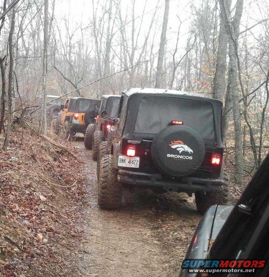 Several Jeeps driving on a muddy, wooded trail. One Jeep has a Broncos logo on its spare tire cover