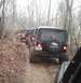 1987 Ford Bronco - A line of off-road vehicles navigates a narrow, dirt path through a forest during the day.